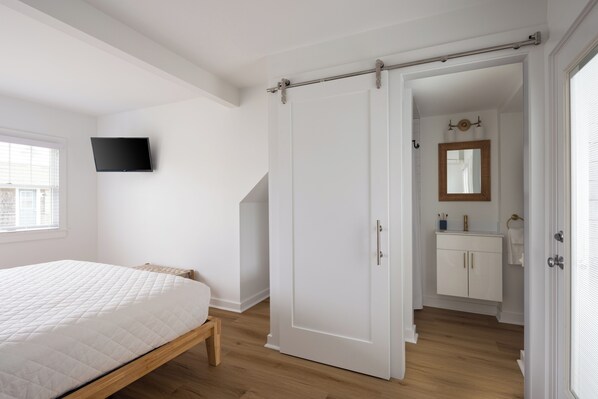 Un dormitorio con cama, una puerta corredera, un televisor montado en la pared y un baño visible a través de una puerta abierta.