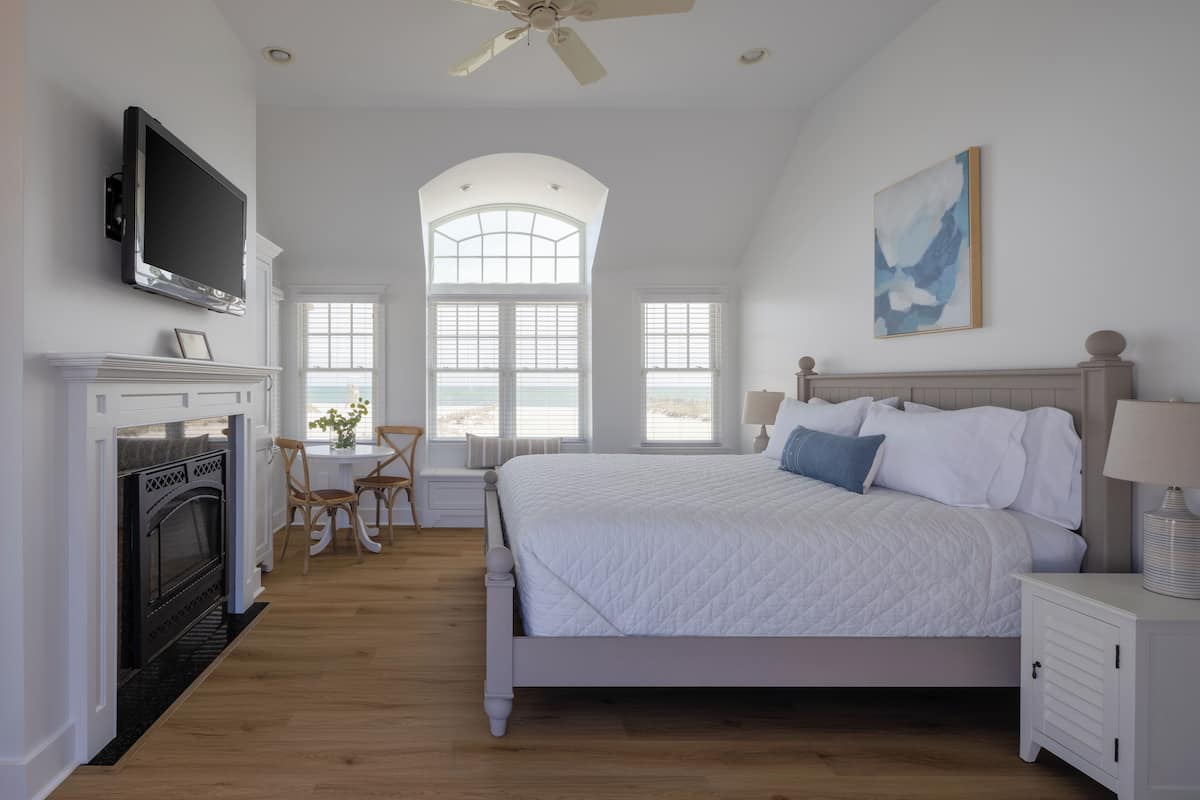 A bedroom with a large bed, a fireplace, a television, and a view of the outdoors through large windows.