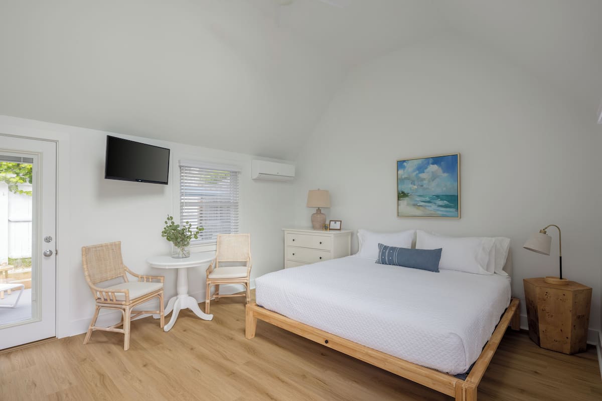 A bedroom with a wooden bed, white walls, a flat-screen TV, a small table with chairs, a nightstand, and a painting of a beach scene.