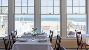 A dining area with a table set for two, large windows with a beach view, and wooden chairs. - Inn on the Beach  (Harwich Port)