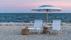 Two white lounge chairs, a beach umbrella, and a wicker basket on a sandy beach with the ocean in the background. - Inn on the Beach  (Harwich Port)