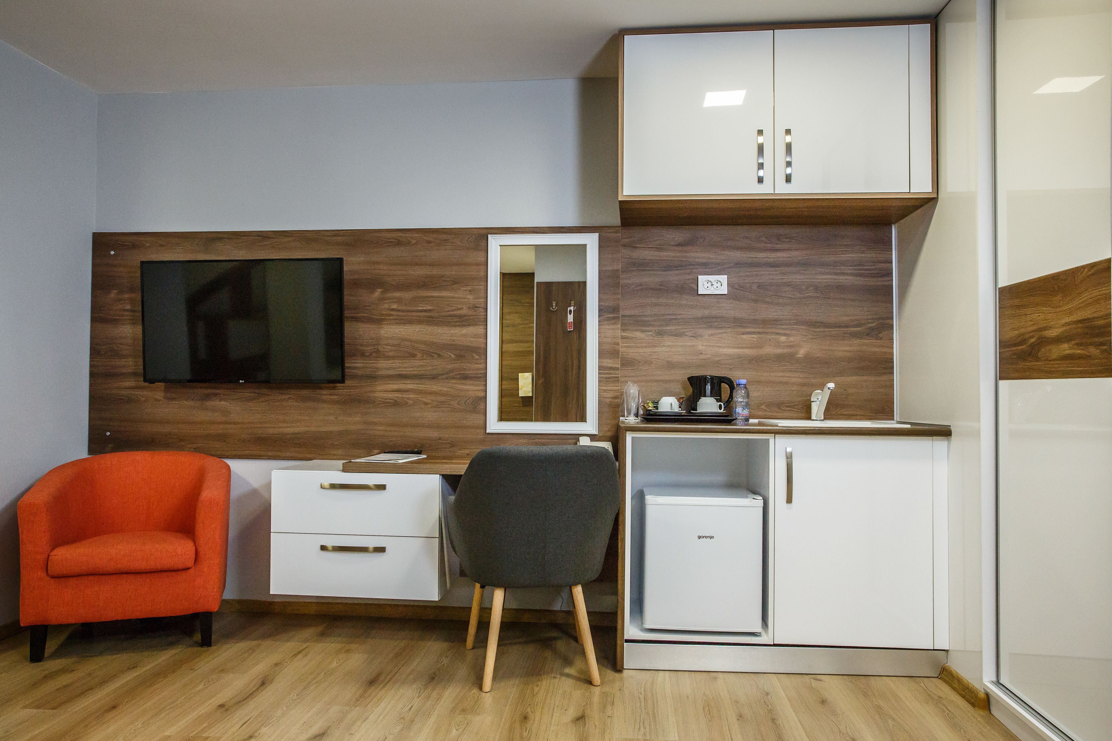 A compact room with a wooden accent wall, a small kitchenette, a desk with drawers, a chair, and a television mounted on the wall.
