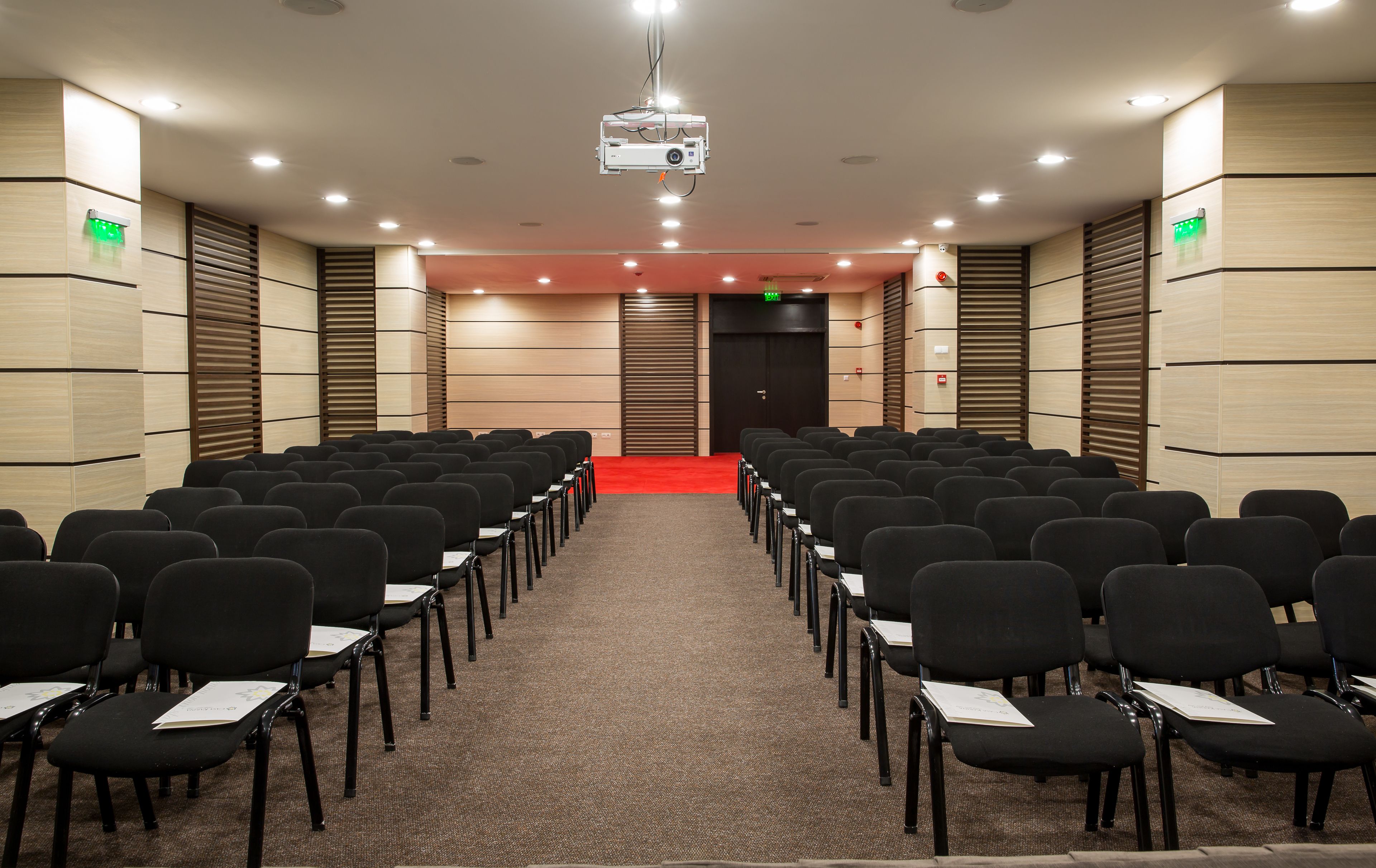 Un salón de conferencias con filas de sillas negras, piso alfombrado y una alfombra roja que lleva a una puerta.