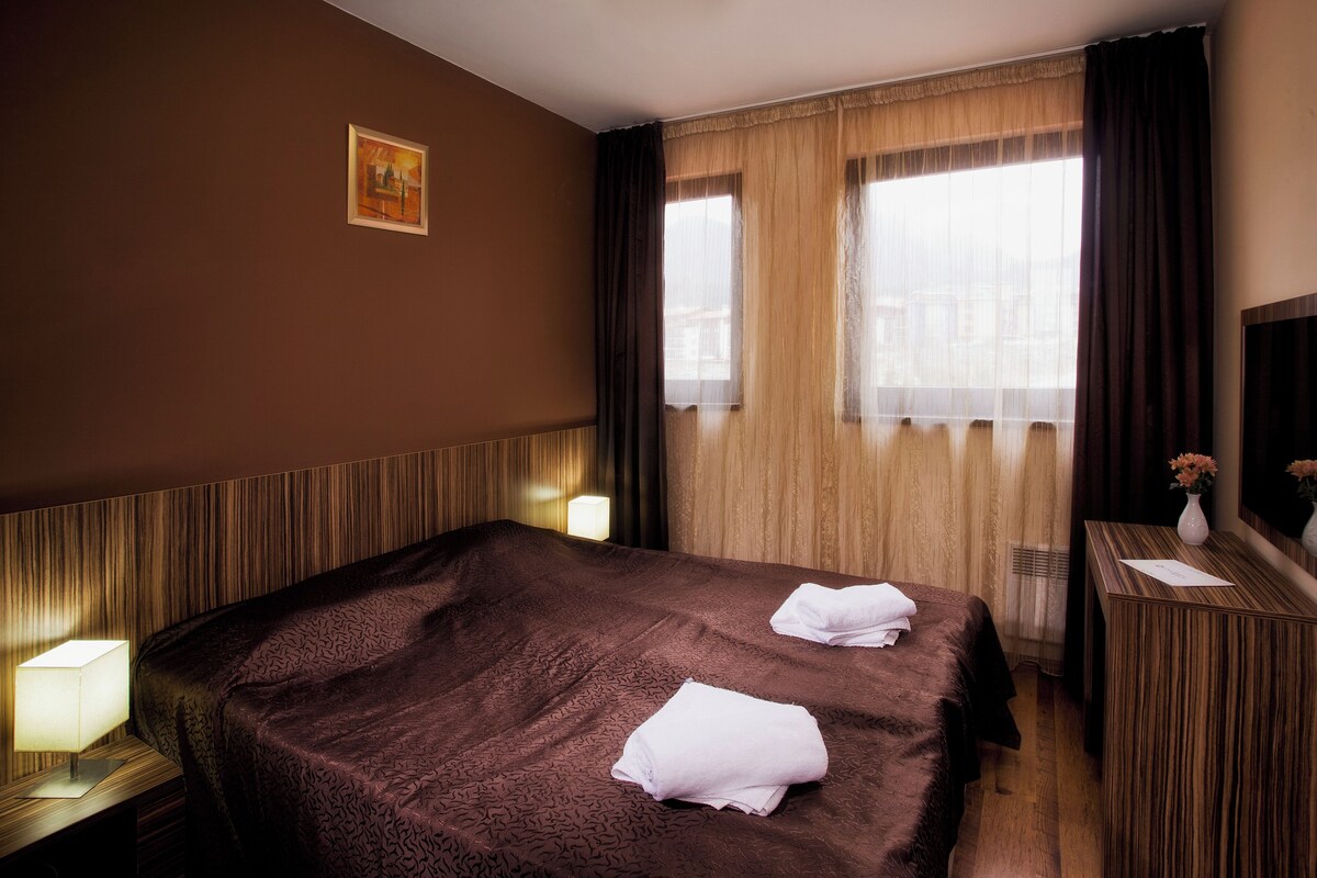 A hotel room with a large bed, a bedside table with a lamp, a mirror, and a window with curtains.