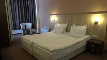 Quarto de hotel com uma cama grande, duas mesas de cabeceira, uma cadeira e uma mesa com garrafa e copo.