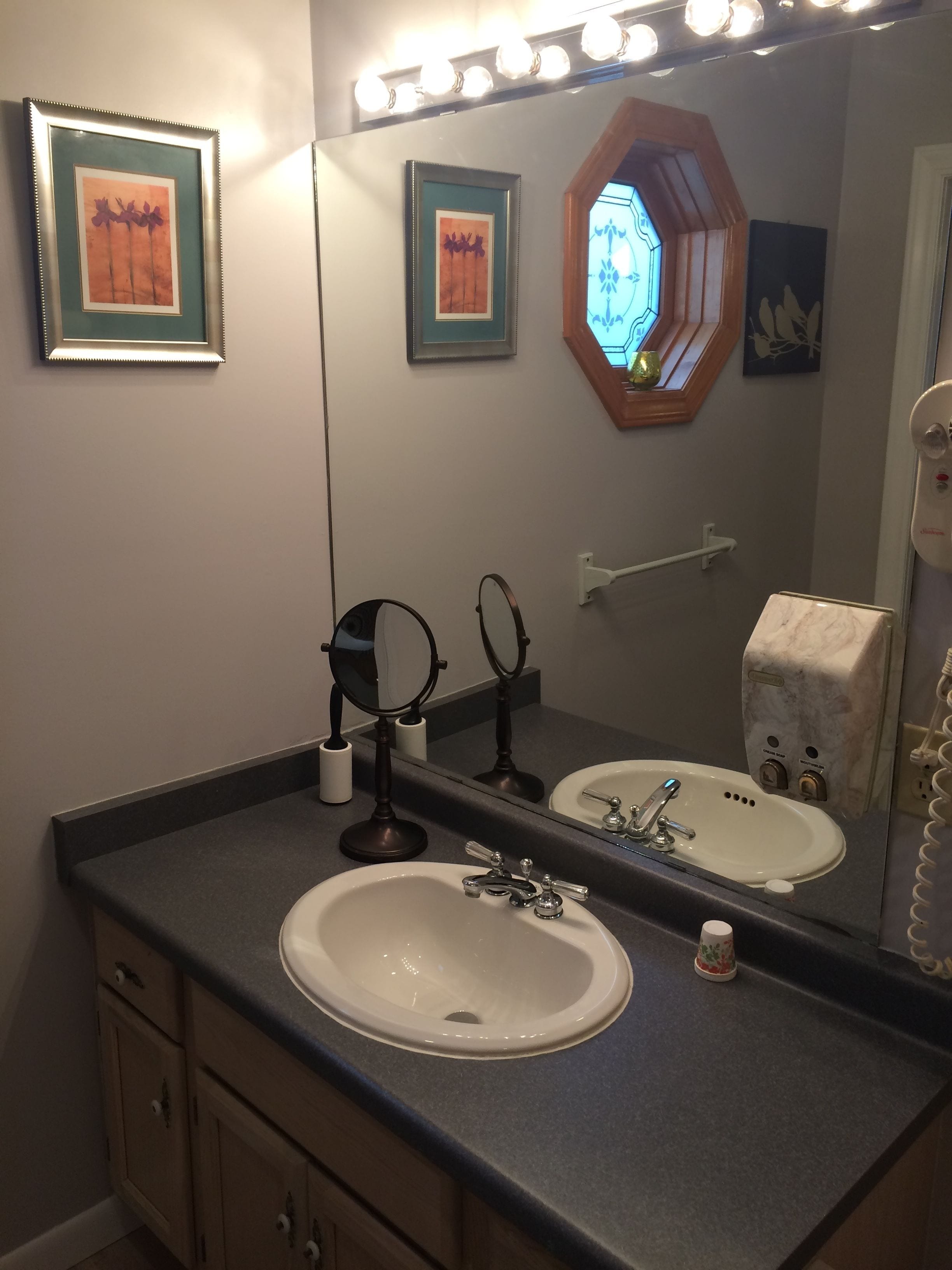 A bathroom with a large mirror, two framed pictures, a sink, a vanity, and a towel dispenser.