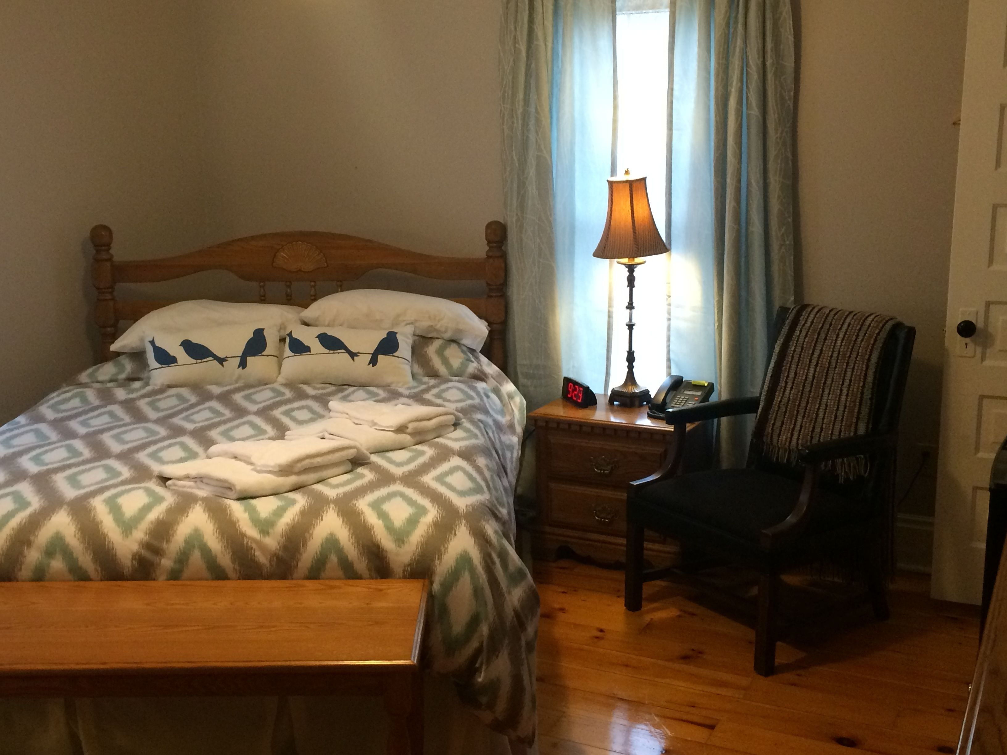 A neatly made bed with patterned bedding, a bedside lamp, a chair, and a wooden nightstand with a clock and phone.