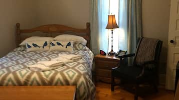 A neatly made bed with patterned bedding, a bedside lamp, a chair, and a wooden nightstand with a clock and phone.