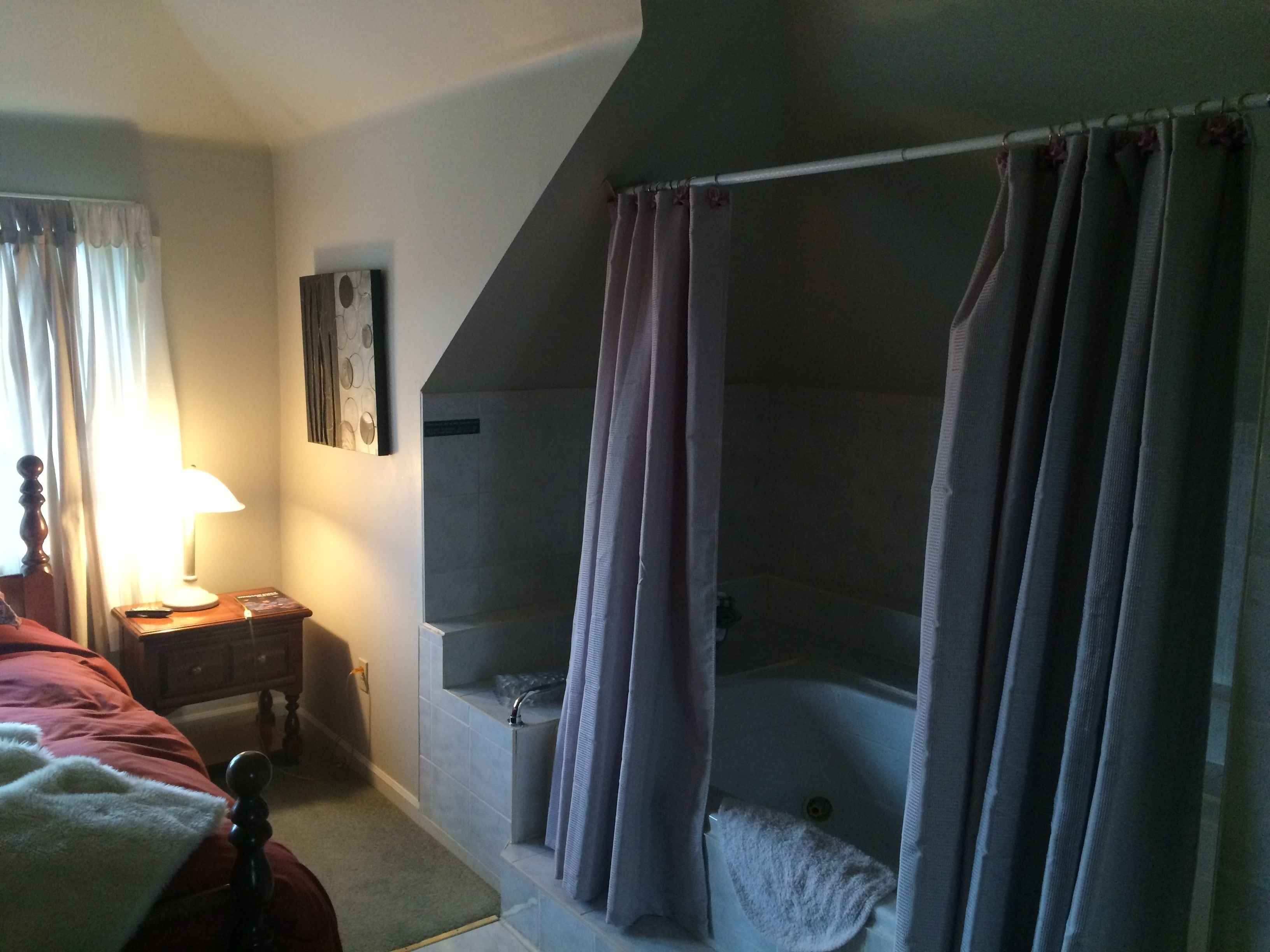 A bedroom with a bed, a bedside table with a lamp, a nightstand, a window with curtains, and a bathroom with a shower and bathtub.