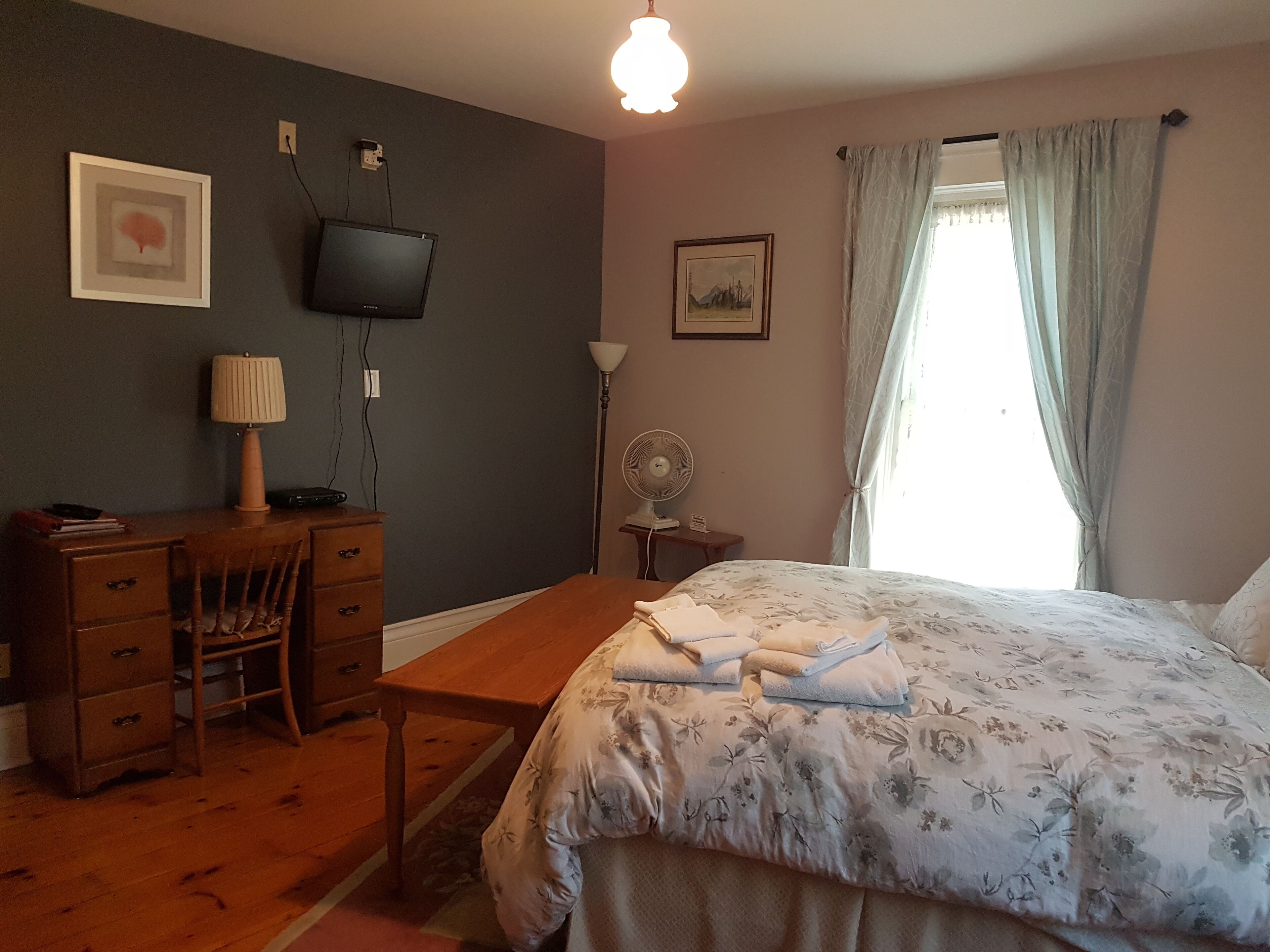 Una camera da letto con un letto, una scrivania, una sedia, una lampada, un televisore, una ventola e una finestra con tende.