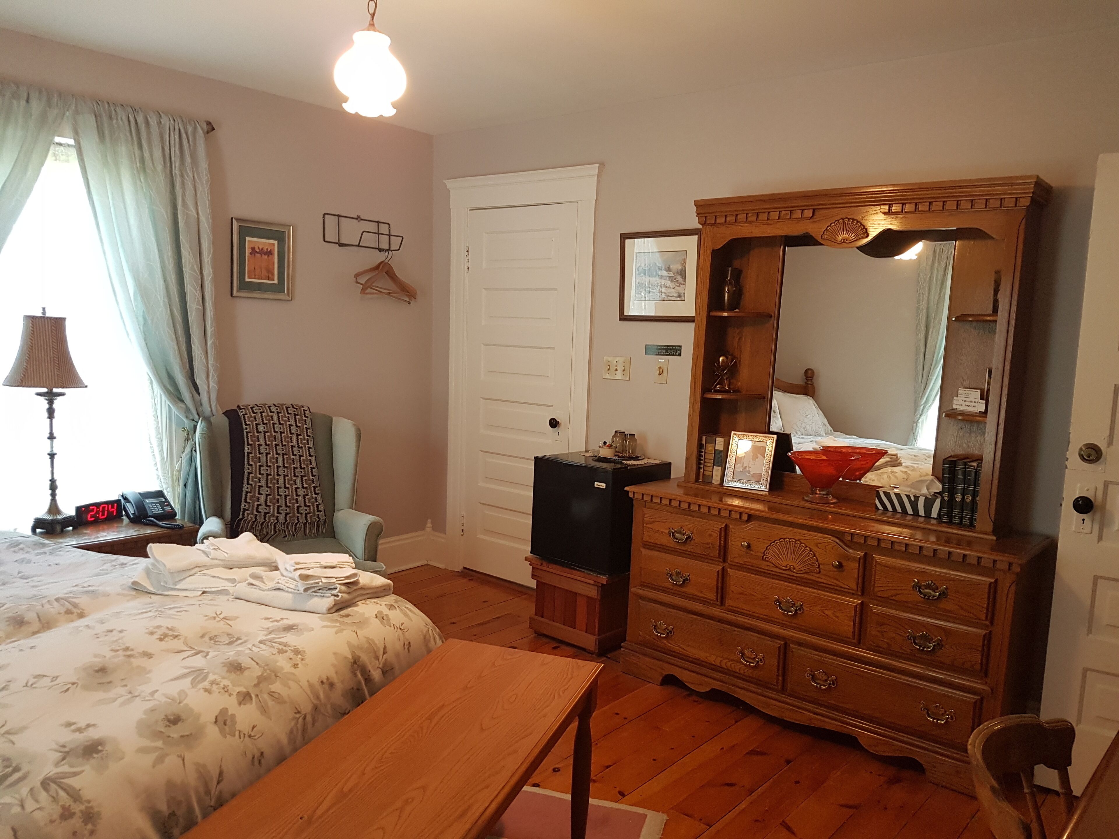 Une chambre à coucher comprenant un lit, une commode, un miroir, une chaise, une table de chevet, une lampe et une horloge.