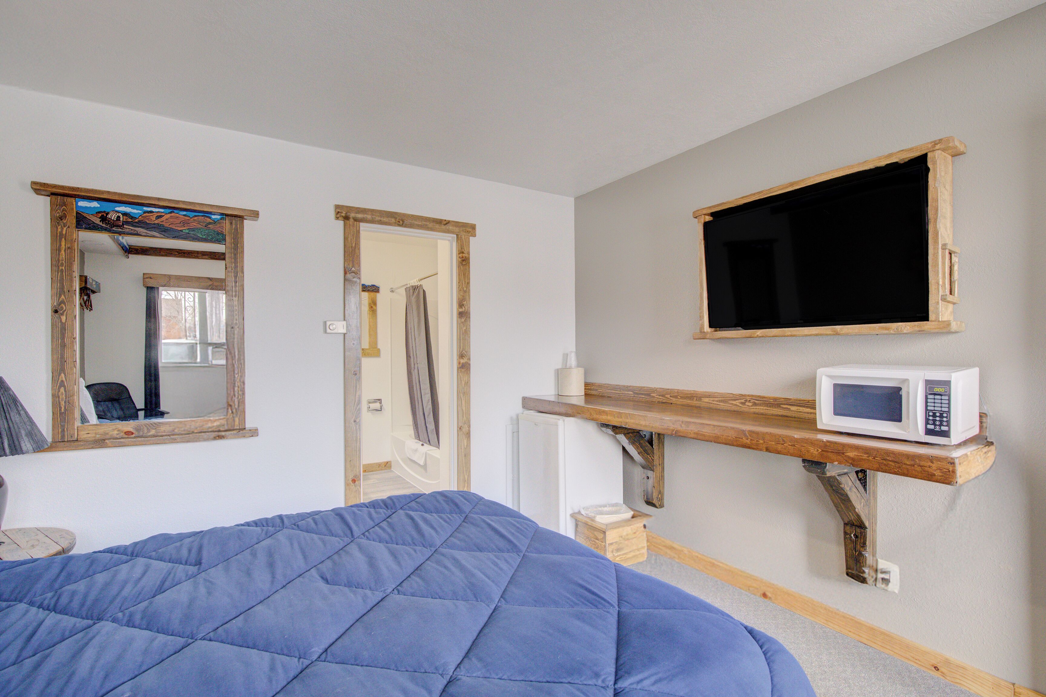 Quarto com uma cama, uma estante de madeira com micro-ondas, um espelho e uma porta que dá para um banheiro.