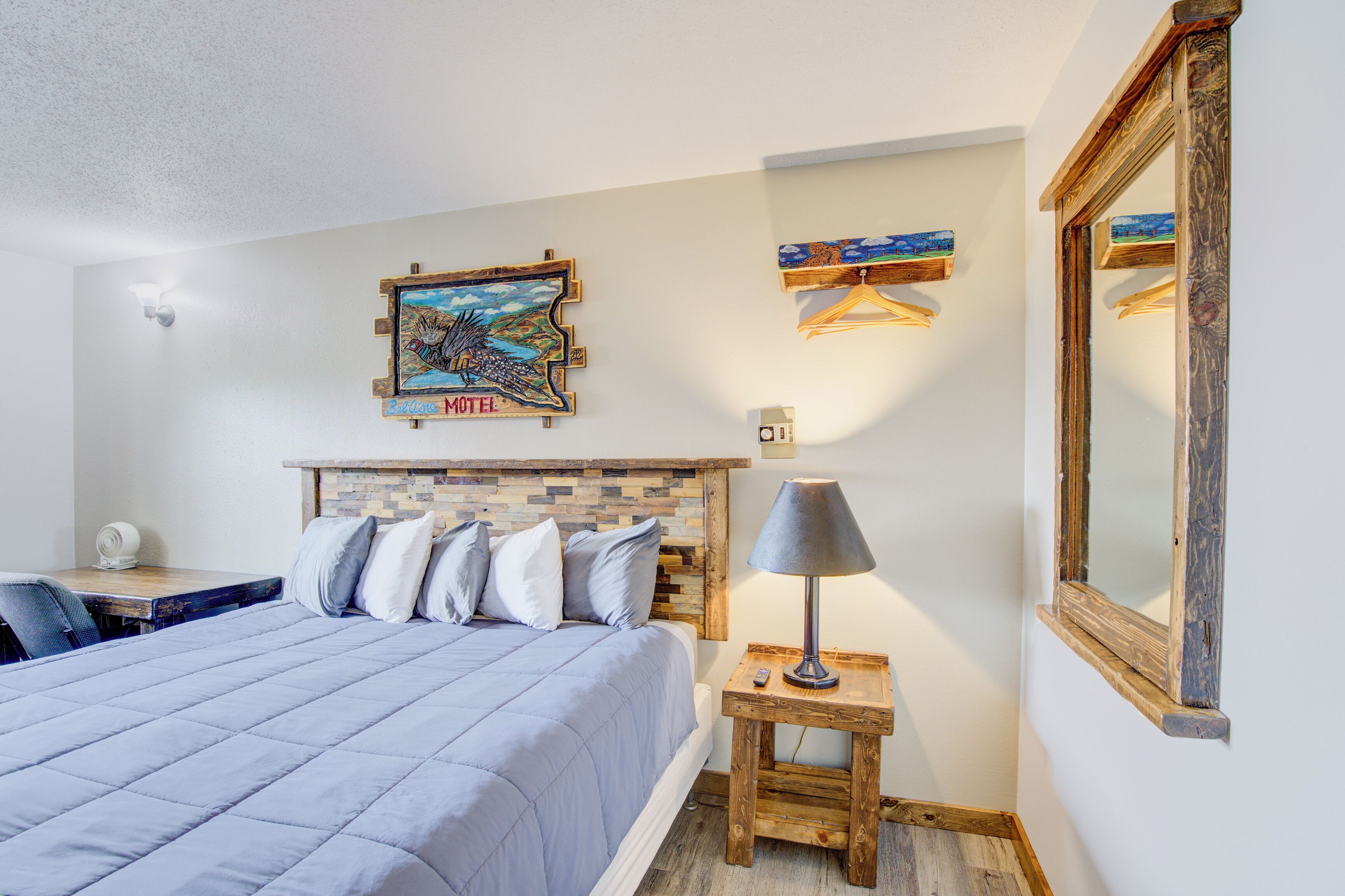 A bedroom with a bed, a desk, a lamp, a mirror, and a wall-mounted painting.