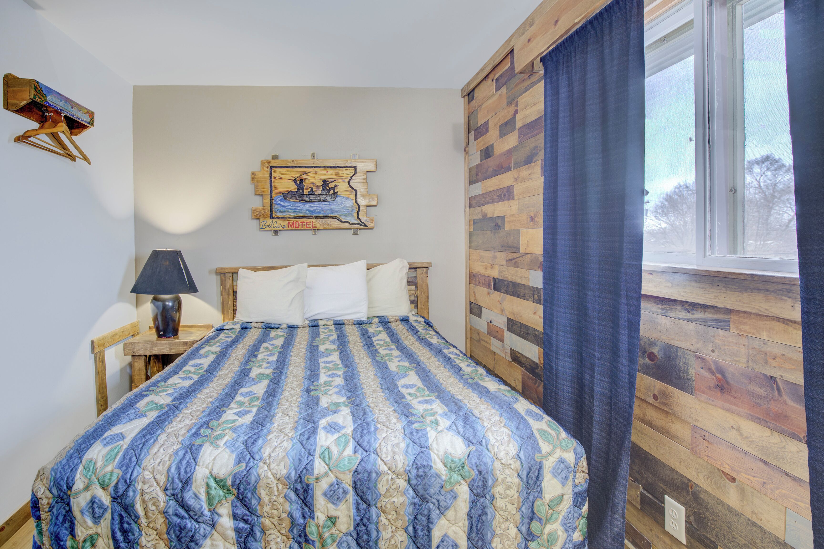 Quarto com cama, mesa de cabeceira, luminária, quadro na parede e janela com cortinas.