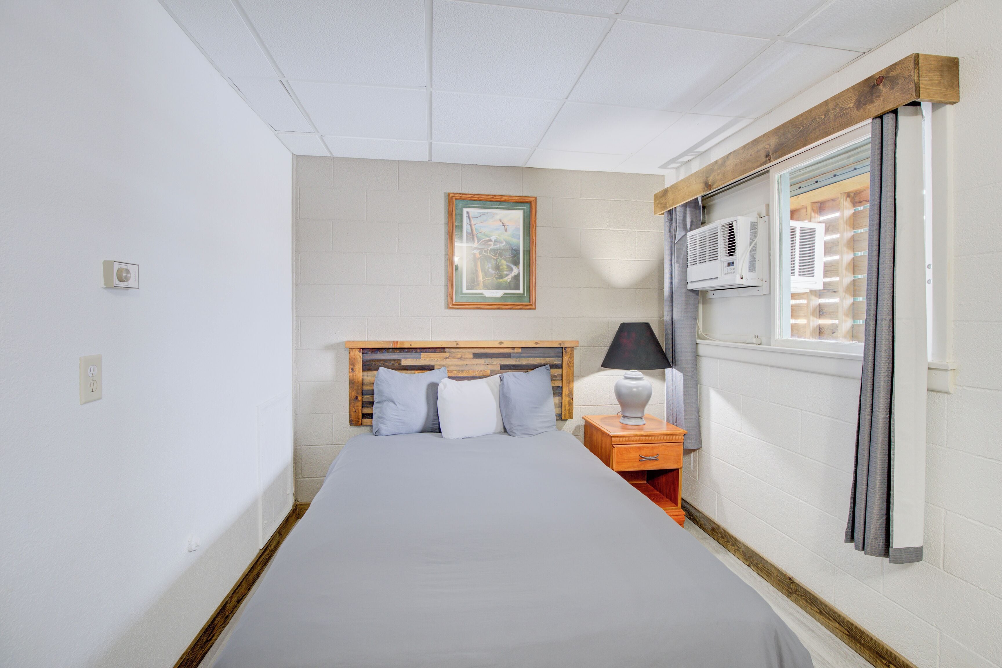 Une chambre à coucher comprenant un lit, une table de chevet, une lampe, un tableau au mur et un appareil de climatisation.