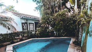 Una piscina con borde de piedra, rodeada de abundante vegetación y un edificio con tejado de baldosas.