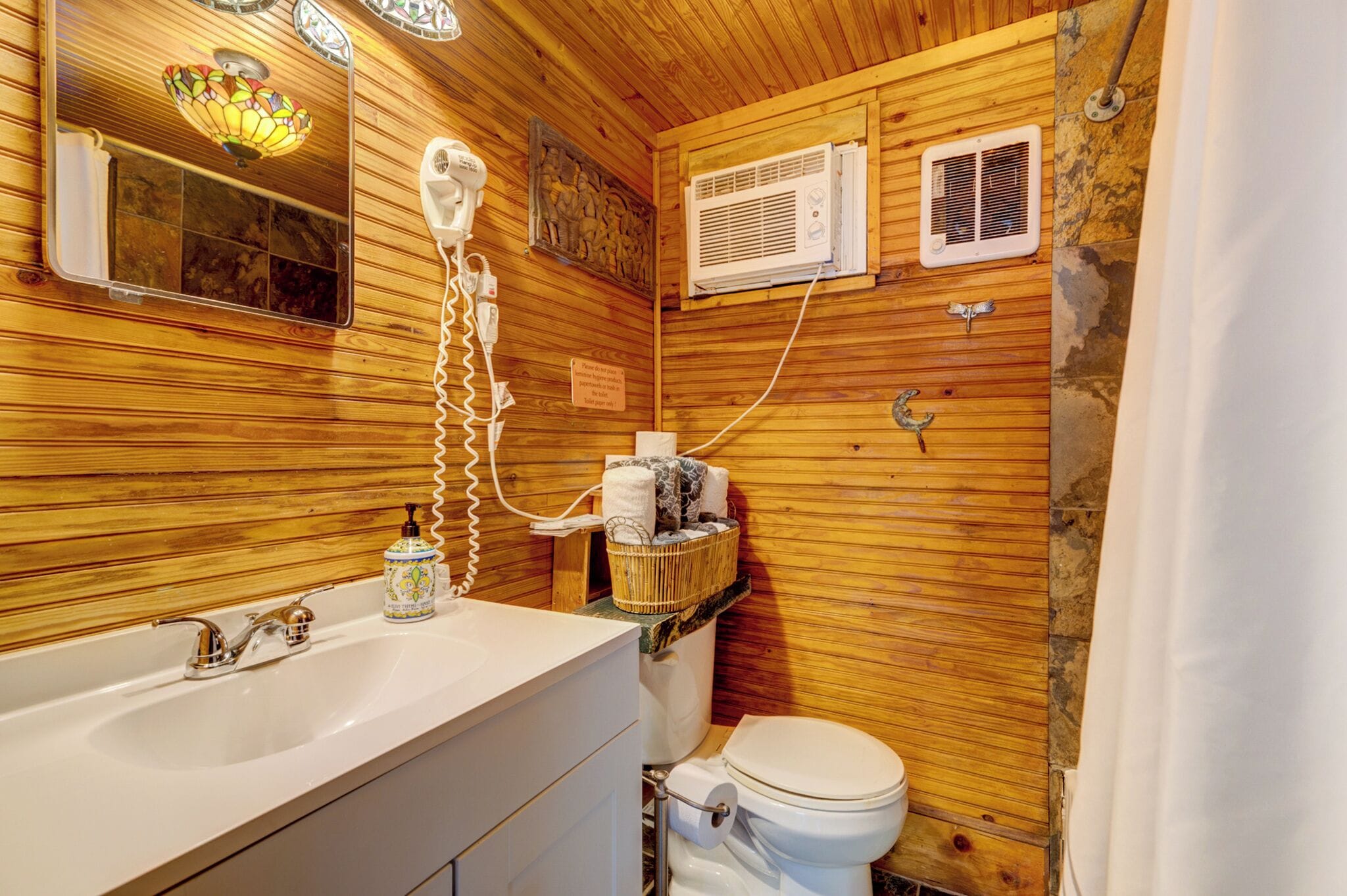 Een houten badkamer in een chalet, met een wastafel, toilet en twee wandventilatoren.