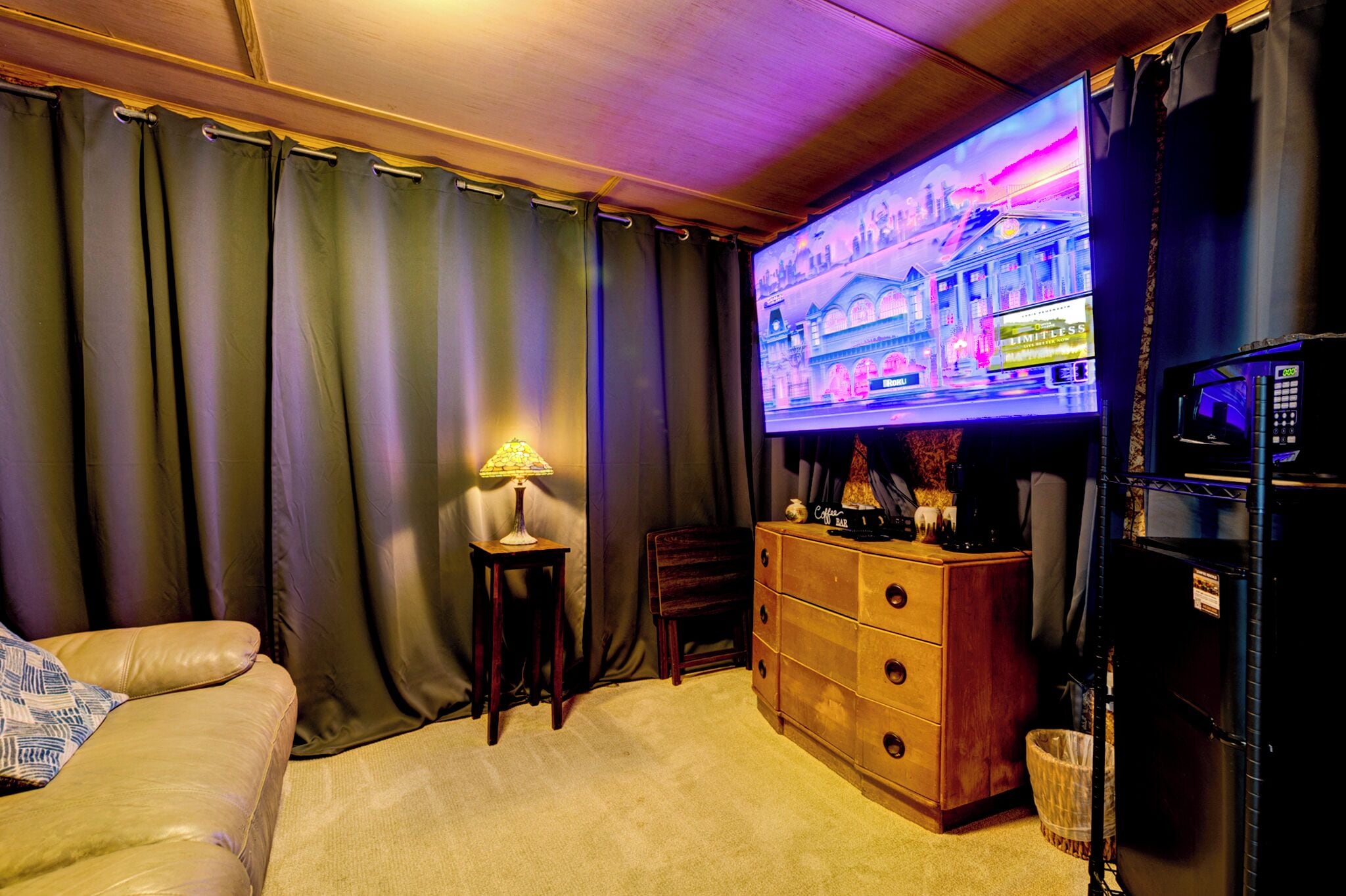 Een knus ingerichte kamer met een groot televisiescherm waarop een spel draait, een houten dressoir, een lamp en een bank.