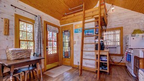 A wooden cabin interior with a dining table, chairs, a ladder, a refrigerator, and a window with a view of trees. - Beachside Boutique Inn – Folly Beach Rooms & Private Cottages (Folly Beach)