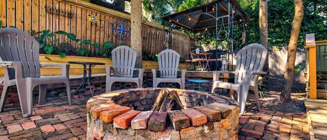 A patio with a brick fire pit, white chairs, and a wooden fence.