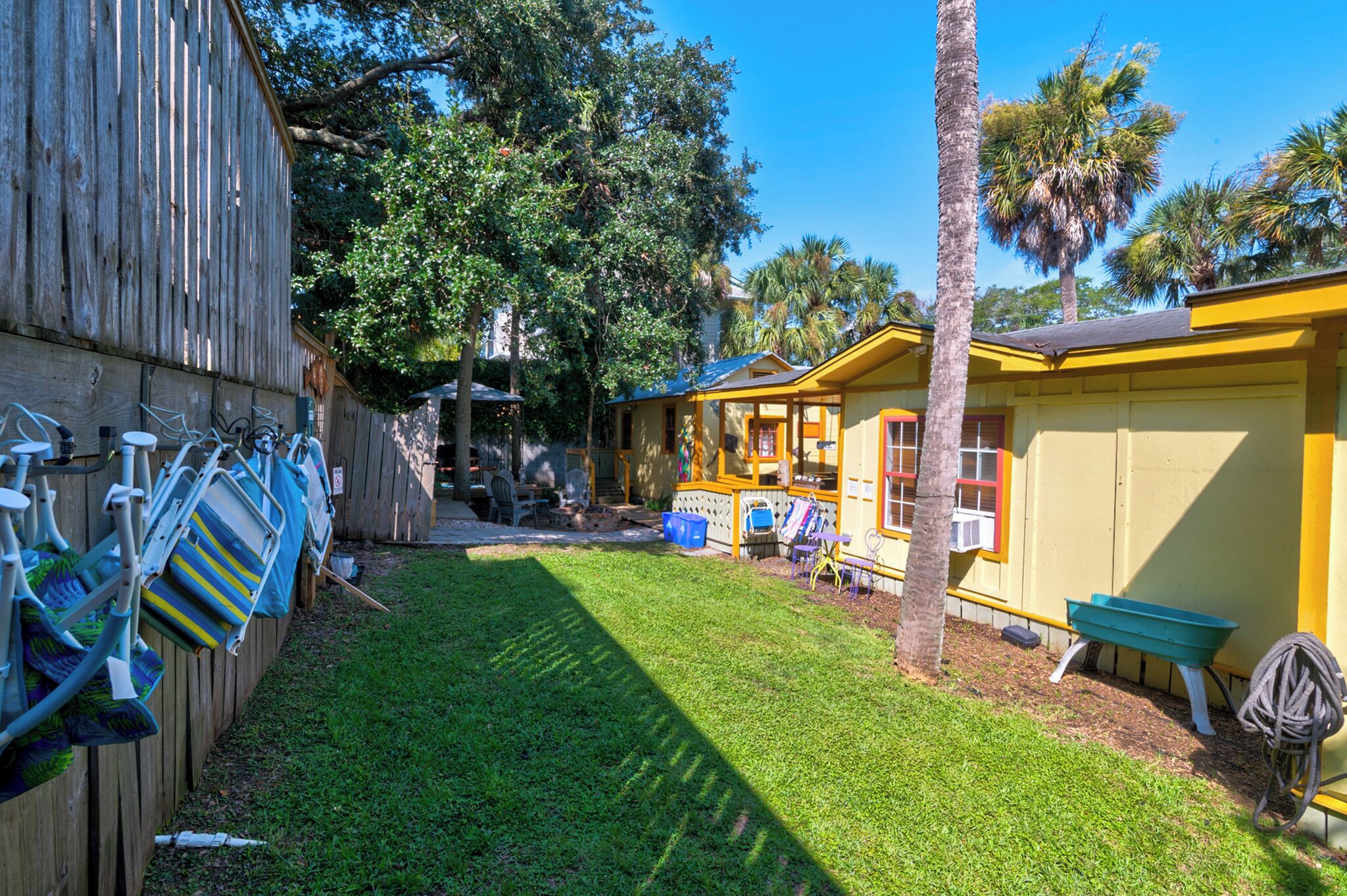 A fenced backyard with a small yellow building, a palm tree, and a green wheelbarrow.
