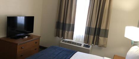 A hotel room with a bed, a TV on a wooden dresser, an air conditioning unit, and a window with curtains.