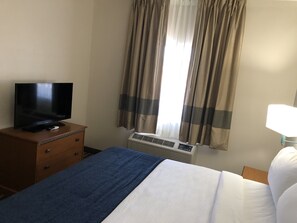 A hotel room with a bed, a TV on a wooden dresser, an air conditioning unit, and a window with curtains.
