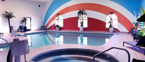 An indoor swimming pool with a colorful mural, lounge chairs, and potted plants.