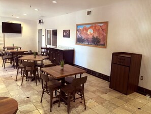 A restaurant with wooden tables and chairs, a kitchen area, and a painting on the wall.