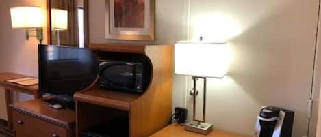 Habitación de hotel con un escritorio de madera, televisor, microondas, cafetera y lámpara.