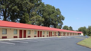 Un motel avec un toit rouge et des murs de couleur beige, composé d’une rangée de chambres de plain-pied dotées de portes et de fenêtres rouges, d’un parking avec des places délimitées et d’arbres en arrière-plan.