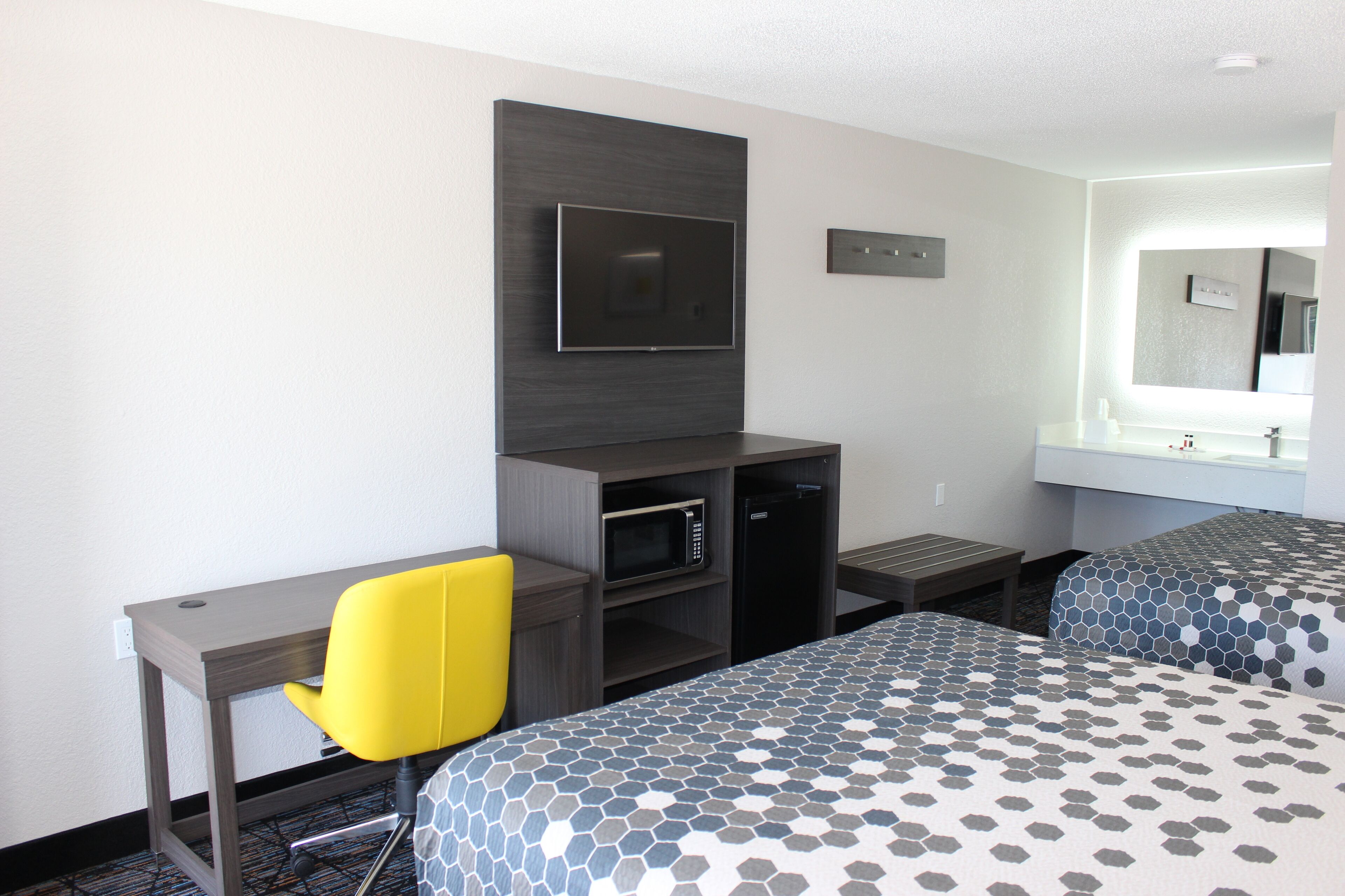 Habitación de hotel con escritorio, televisión, microondas, una mesita pequeña, dos camas con ropa de cama estampada y un espejo grande sobre el lavamanos.