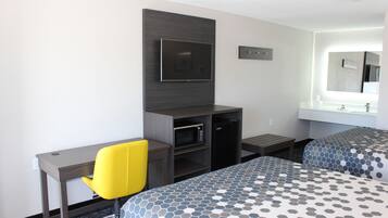 Habitación de hotel con escritorio, televisión, microondas, una mesita pequeña, dos camas con ropa de cama estampada y un espejo grande sobre el lavamanos.