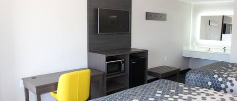 A hotel room with a desk, a TV, a microwave, a small table, two beds with patterned bedding, and a large mirror above the sink.