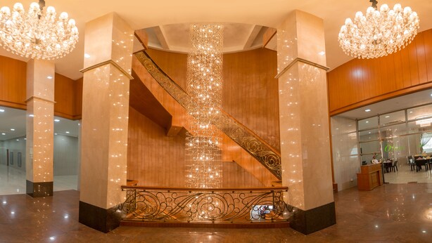 A grand staircase with ornate railings and a chandelier, leading to a lobby area with wooden paneling and a reception desk. - Sky Star Hotel (Yangon)