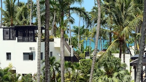 A modern building with a white facade, surrounded by tall palm trees and a clear blue sky. - Aligio Aparthotel & Spa (Las Terrenas)