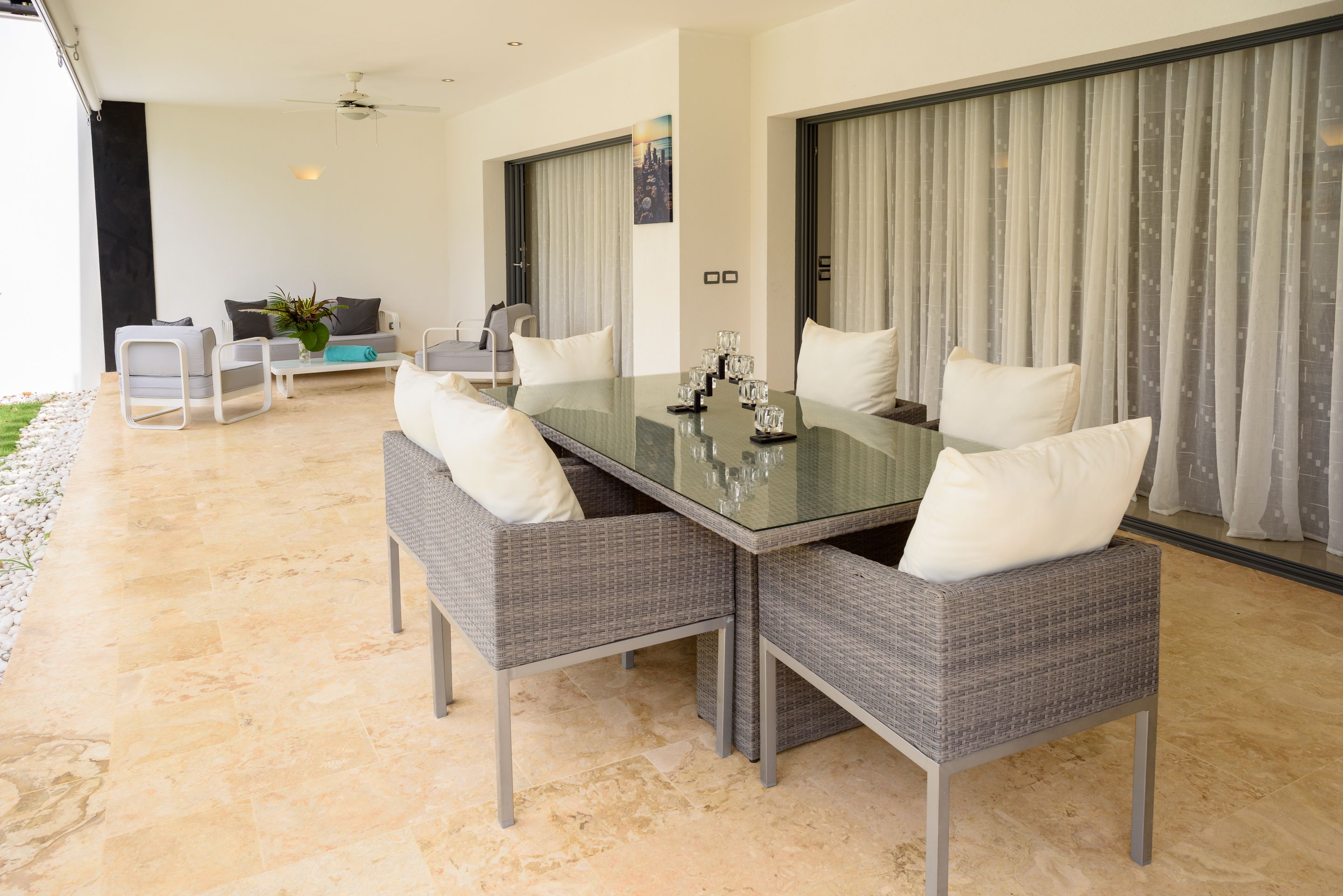 Un coin repas avec une table en verre, des chaises en osier grises et des coussins blancs.