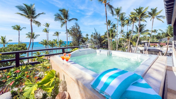 A poolside area with a view of the ocean, surrounded by palm trees and a lounge chair with a striped towel. - Aligio Aparthotel & Spa (Las Terrenas)