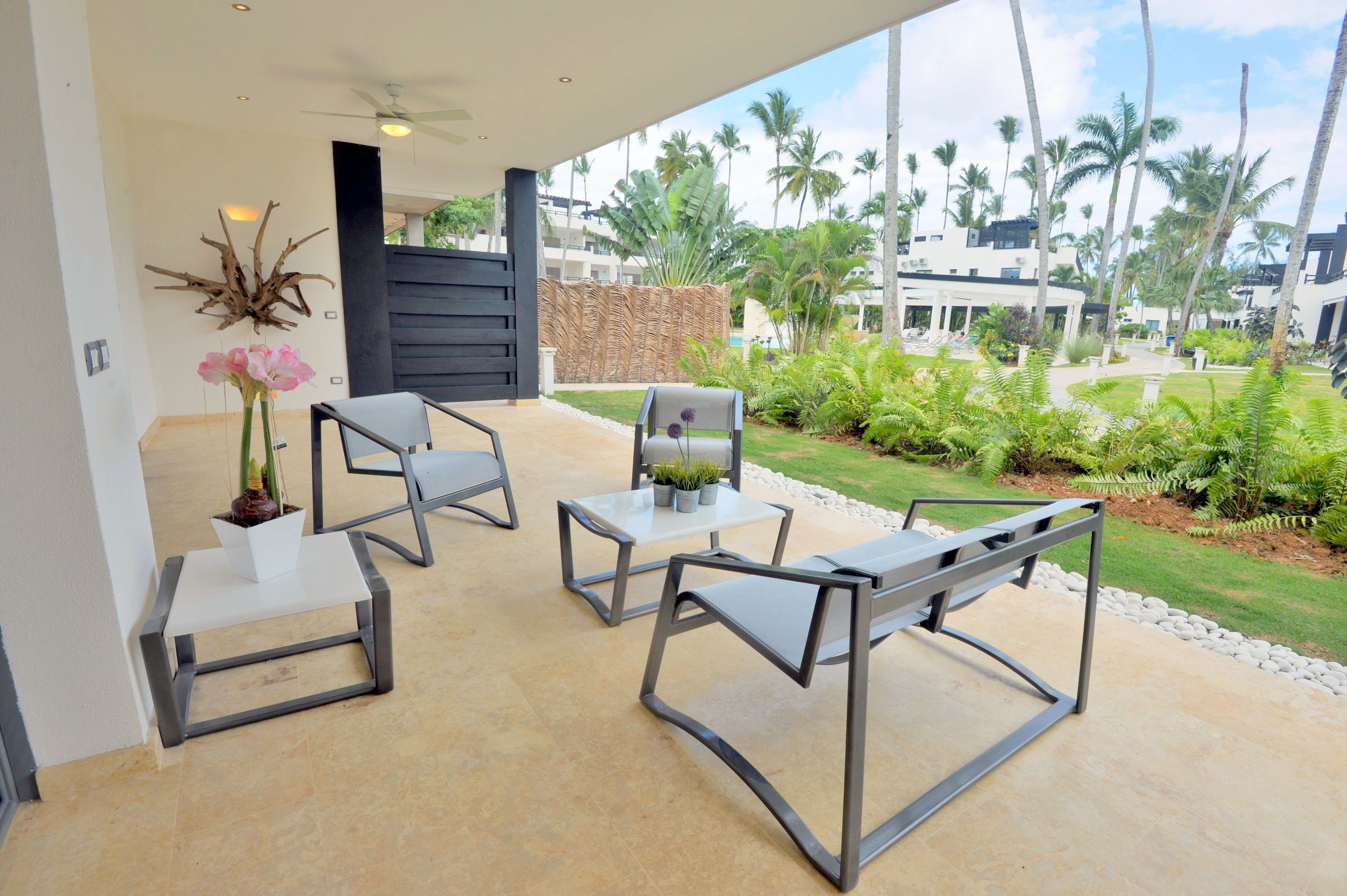 An outdoor patio with modern furniture, a decorative wall piece, and a view of a tropical garden with palm trees.