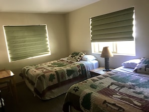 A bedroom with two beds, a nightstand, a lamp, and two windows with blinds. - Twin Peaks Lodge and RV Park (Seward)