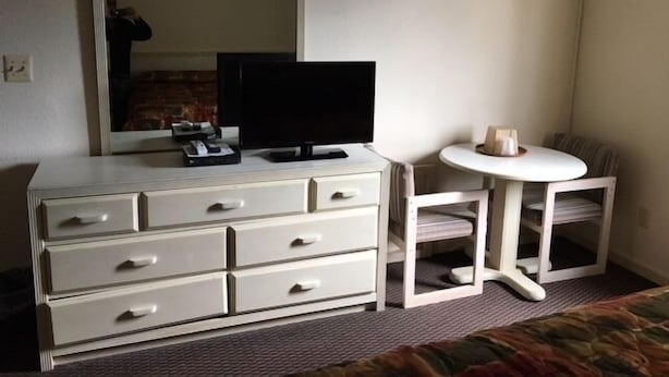 A hotel room with a white dresser, a television, a small round table, and a chair. - Colonade Motel (Hightstown)