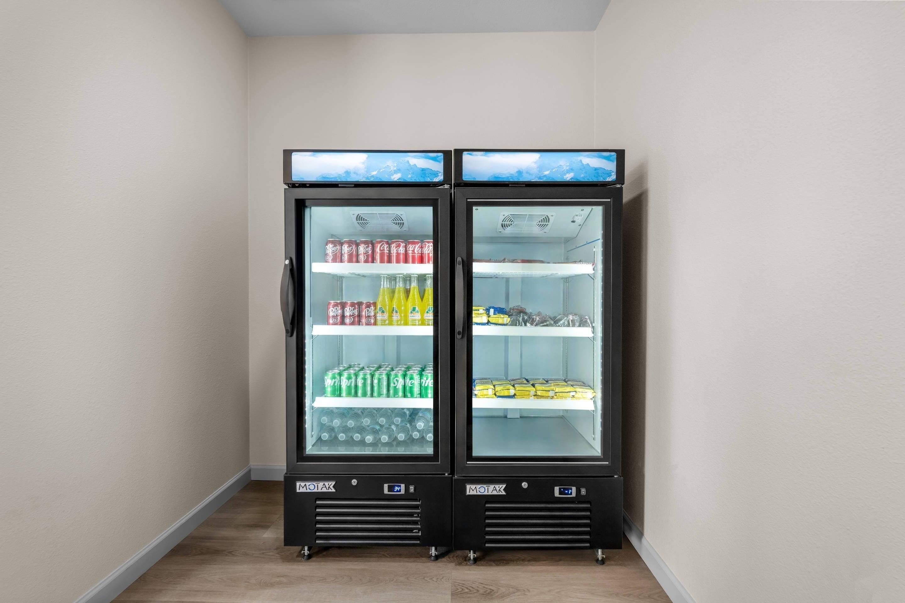 Zwei schwarze Getränke- und Snackautomaten mit Glastüren stehen in einer Ecke mit schlichter Wand und Holzboden.