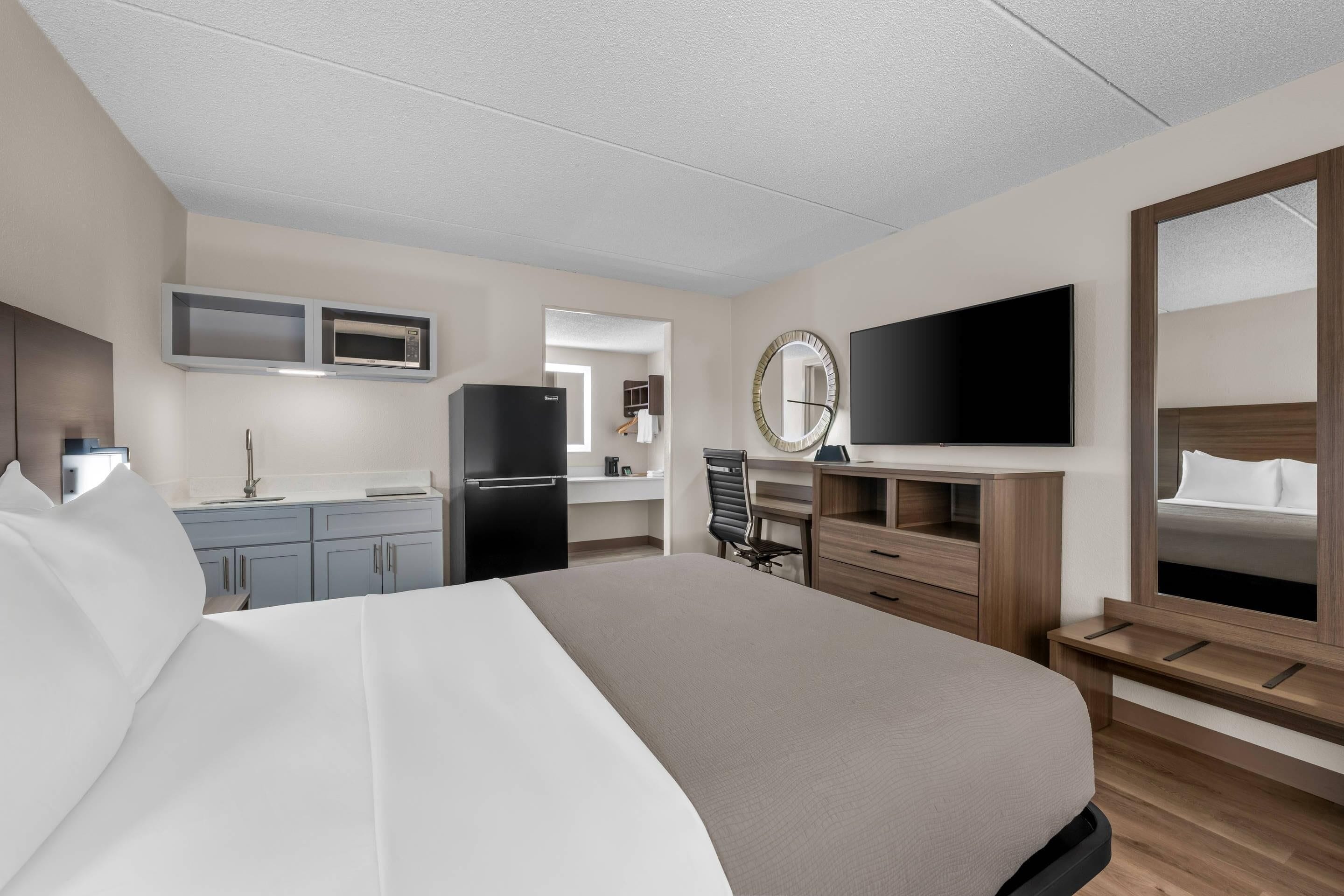 A hotel room with a bed, a desk with a chair, a TV, a microwave, a refrigerator, and a mirror.