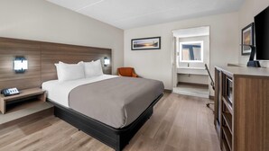 A hotel room with a bed, bedside table, chair, desk, TV, and artwork on the walls.