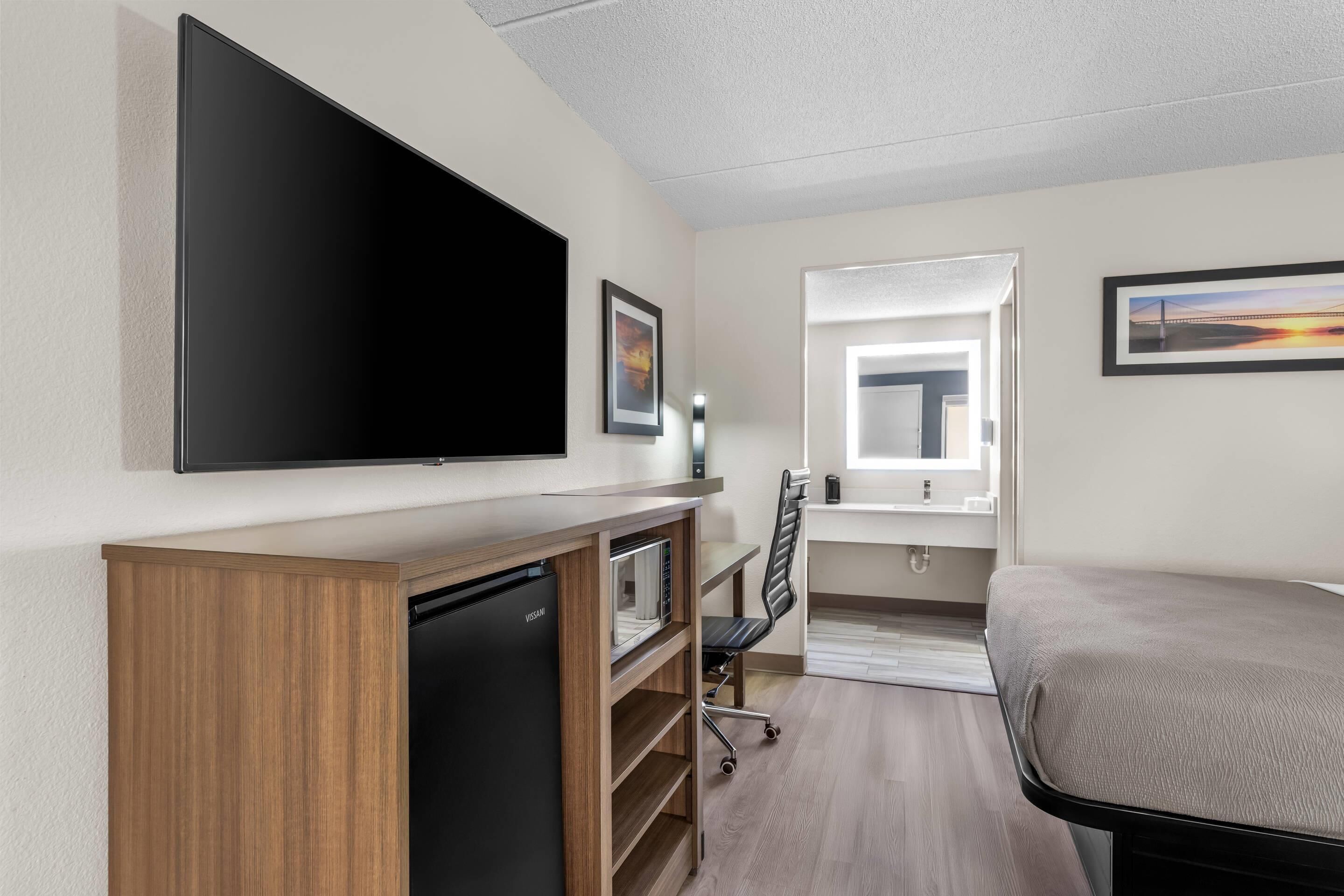 A hotel room with a flat-screen TV, a desk with a microwave, a chair, a bed, and a window with a view.