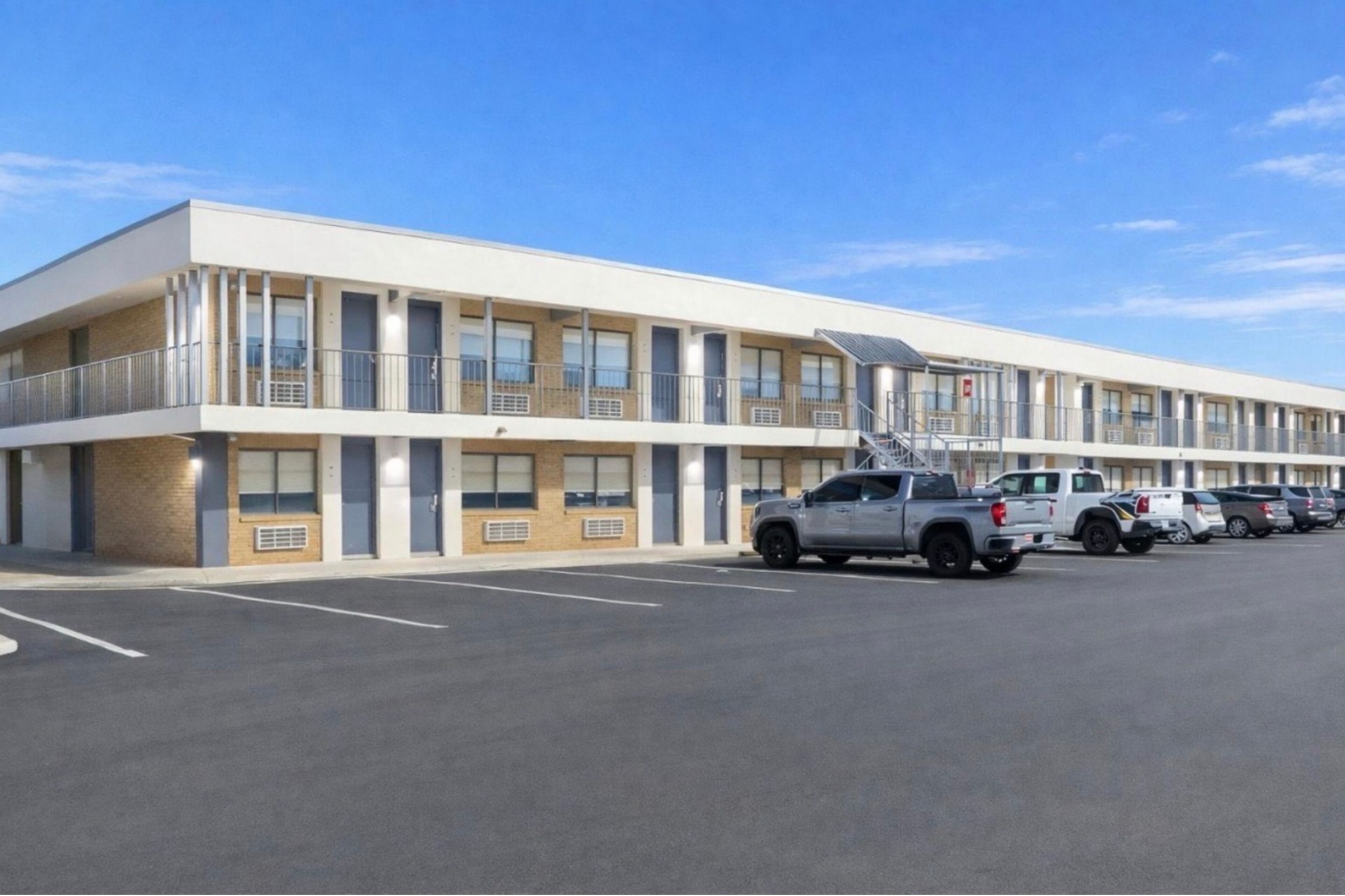 Un motel con una hilera de habitaciones, un estacionamiento y algunos vehículos estacionados.