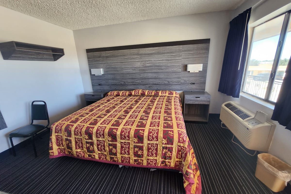 Ein Hotelzimmer mit einem großen Bett, einem Schreibtisch, einem Sessel, einem Fenster mit Vorhängen und einer Wand-Klimaanlage.