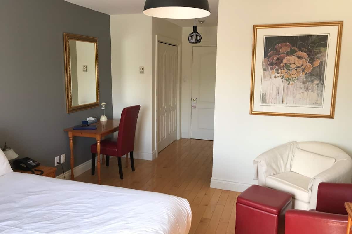 A hotel room with a bed, a desk with a chair, a white armchair, a painting, and a large pendant light.