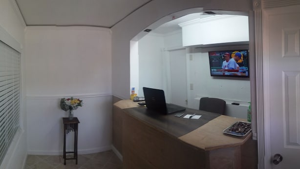 Ein Empfangstresen mit einem Laptop, einem kleinen Tisch mit Blumen in einer Vase und einem Fernseher, der ein Sportevent zeigt.