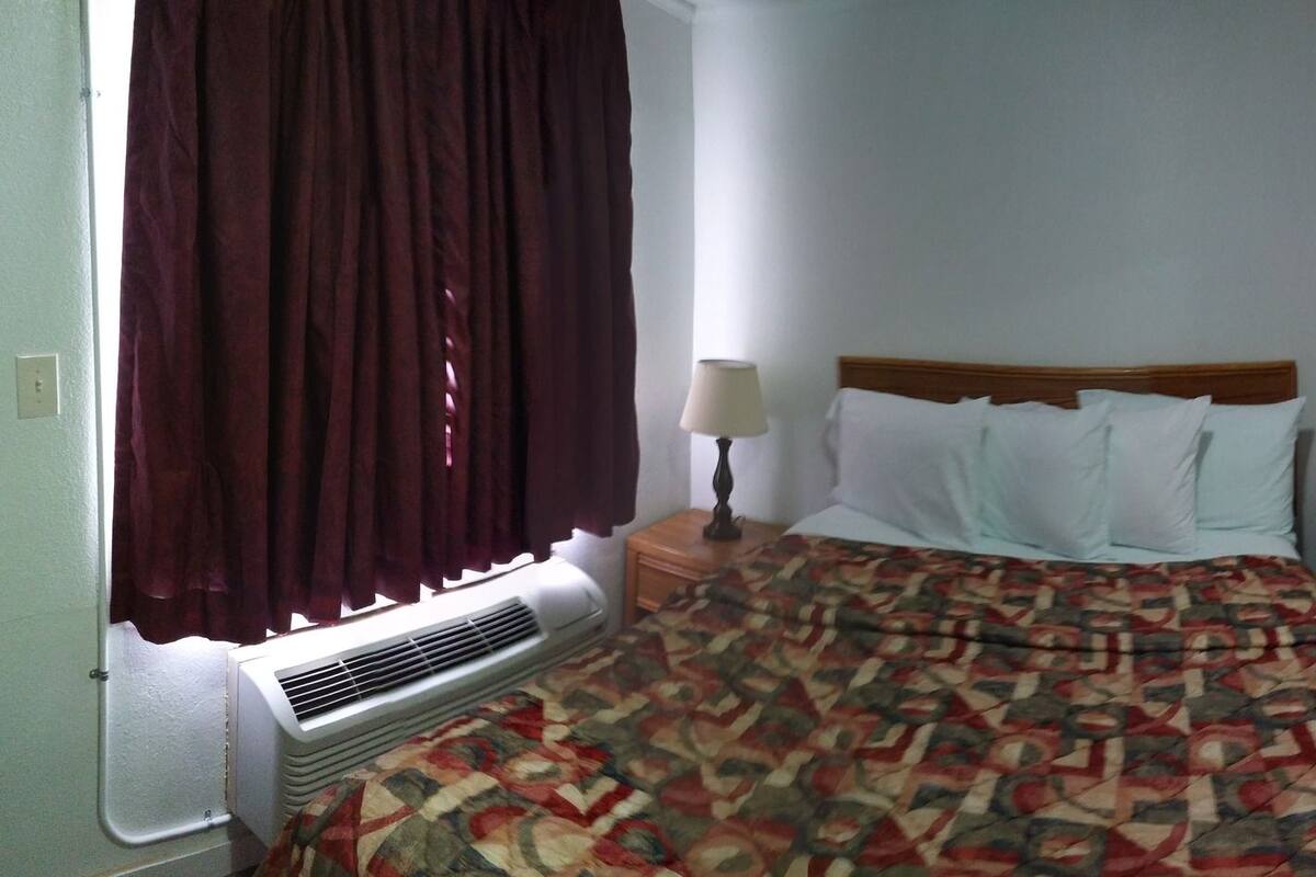 A bedroom with a bed, a bedside table, a mirror, a TV, and a window with curtains.